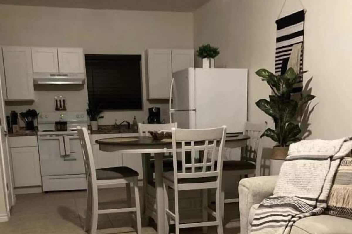Una cocina moderna con armarios blancos, una mesa de comedor y sillas. En la sala de estar, hay un sofá con un cojín estampado.