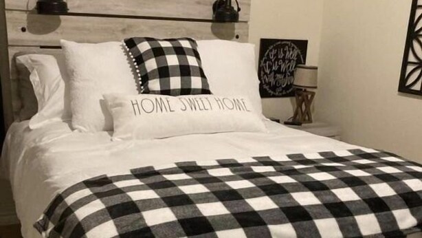Una cama impecable con una manta a cuadros blanco y negro, un cojín con la frase “HOME SWEET HOME” y un cabecero con un acabado de madera clara.