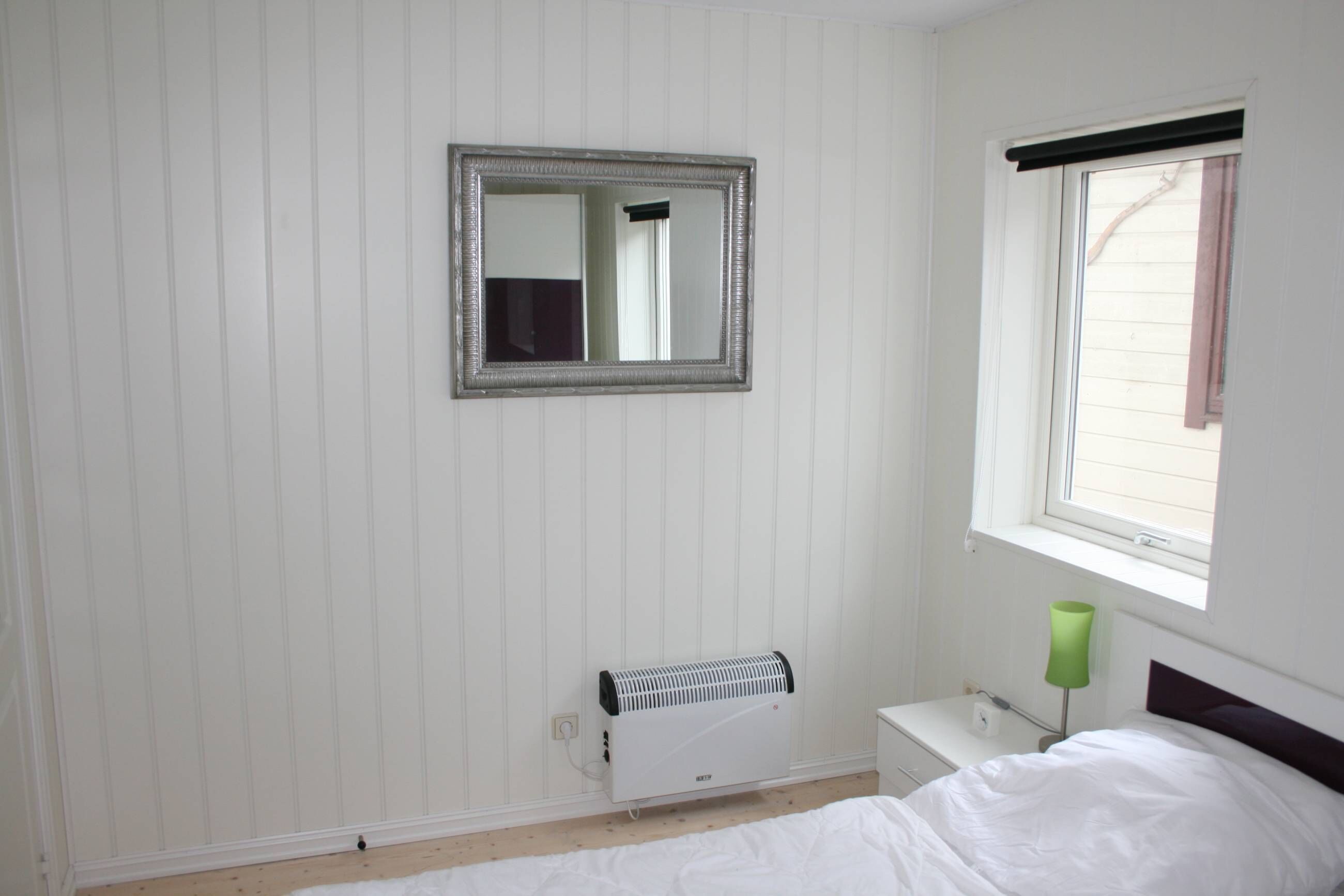 A bedroom with a radiator, a bedside table, a lamp, a bed, and a window with blinds.