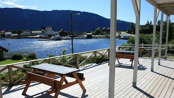 Una terraza de madera con mesas de picnic, con vistas a un cuerpo de agua, barcos y casas al fondo.