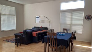 Un salon avec un canapé en cuir noir, une table à manger avec des chaises, un plancher en bois et deux fenêtres équipées de stores.