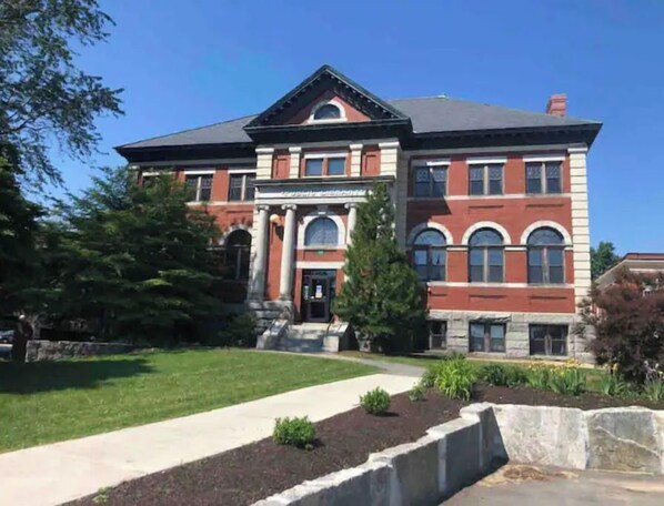 Stately building with a central entrance, columns, and pediment, set amidst a well-manicured lawn and mature trees.