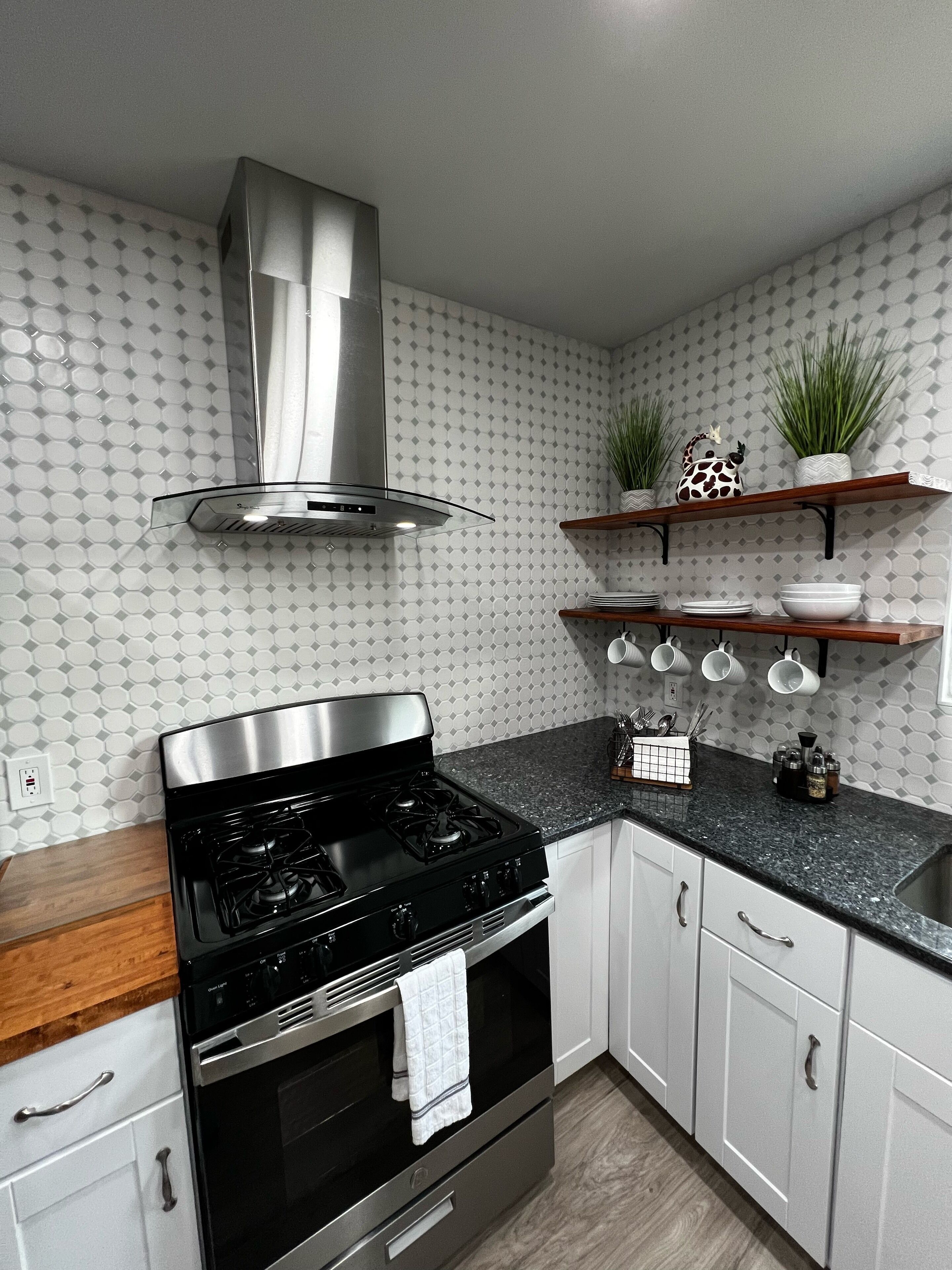 A modern kitchen with a stainless steel stove, oven, and a range hood. There are white cabinets, a granite countertop, and a backsplash with a geometric pattern.