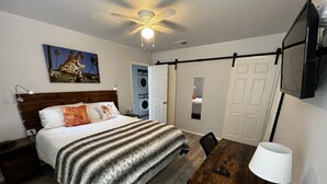 Une chambre à coucher comprenant un lit, un bureau, une chaise, un ventilateur de plafond, une télévision, un tableau et un lave-linge.