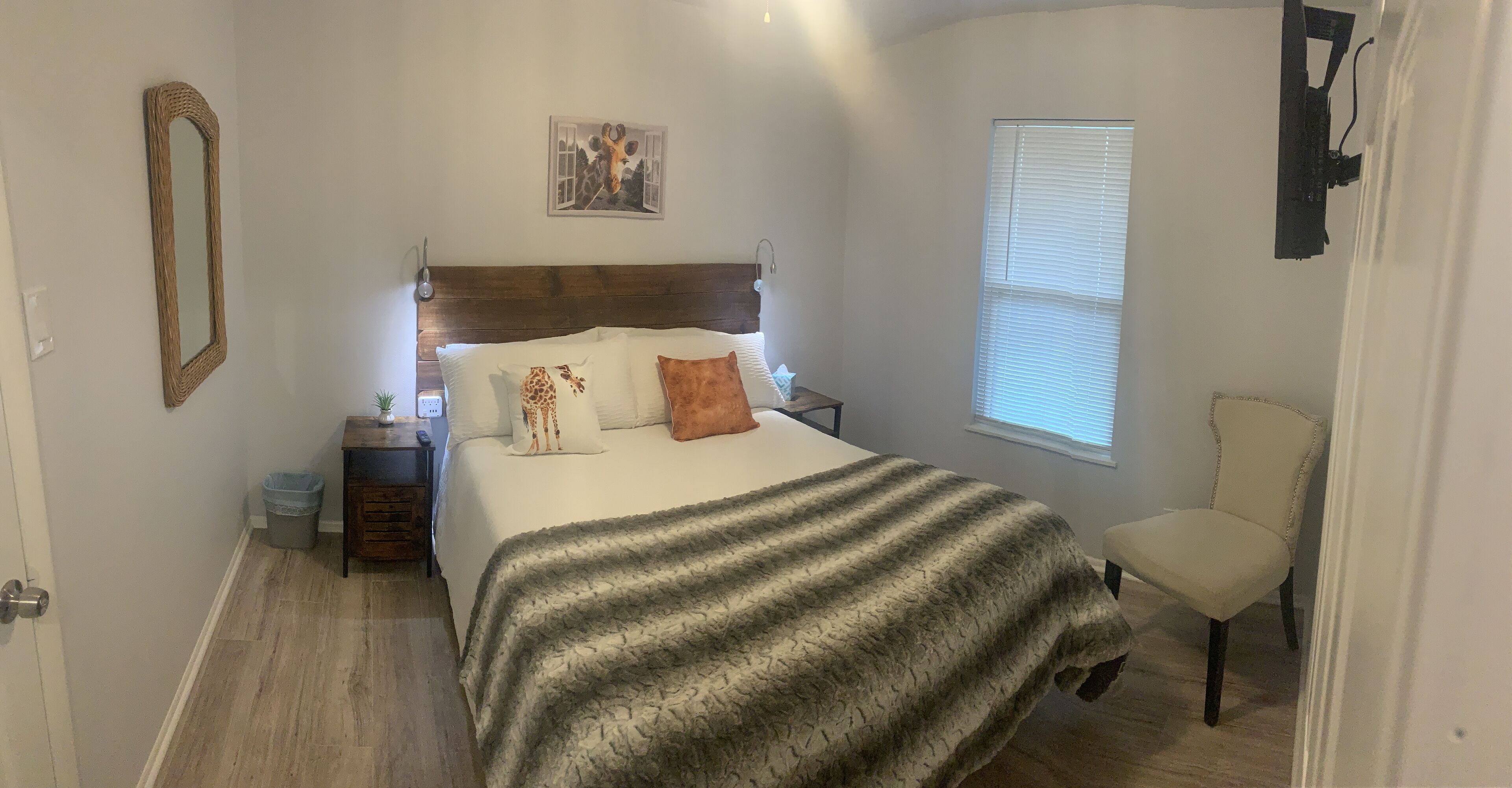 A bedroom with a large bed, a wooden headboard, a nightstand, a chair, a window with blinds, and a wall-mounted mirror.