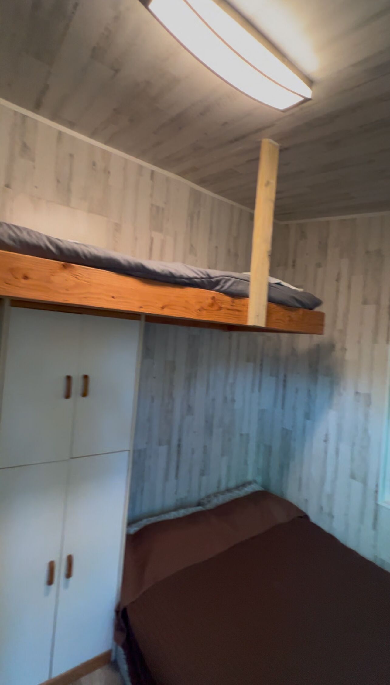 Un lit superposé avec une tête de lit en bois, une armoire à deux portes et une fenêtre donnant sur l’extérieur.