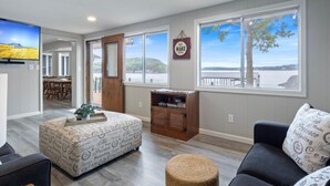 Un vaste salon doté d’une grande fenêtre offrant une vue sur l’eau, un coin repas avec des meubles en bois et un espace salon confortable agrémenté d’un pouf rond.