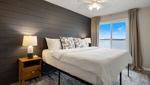 Une chambre à coucher avec un lit, des lampes de chevet, une table de nuit, un ventilateur de plafond et une vue sur une étendue d’eau par la fenêtre.