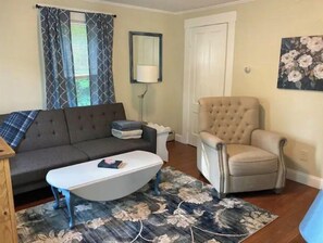 Ein Wohnzimmer mit einem dunkelgrauen Sofa, einem weißen Couchtisch, einem beige farbenen Sessel, einem Blumenteppich und einem Gemälde an der Wand.