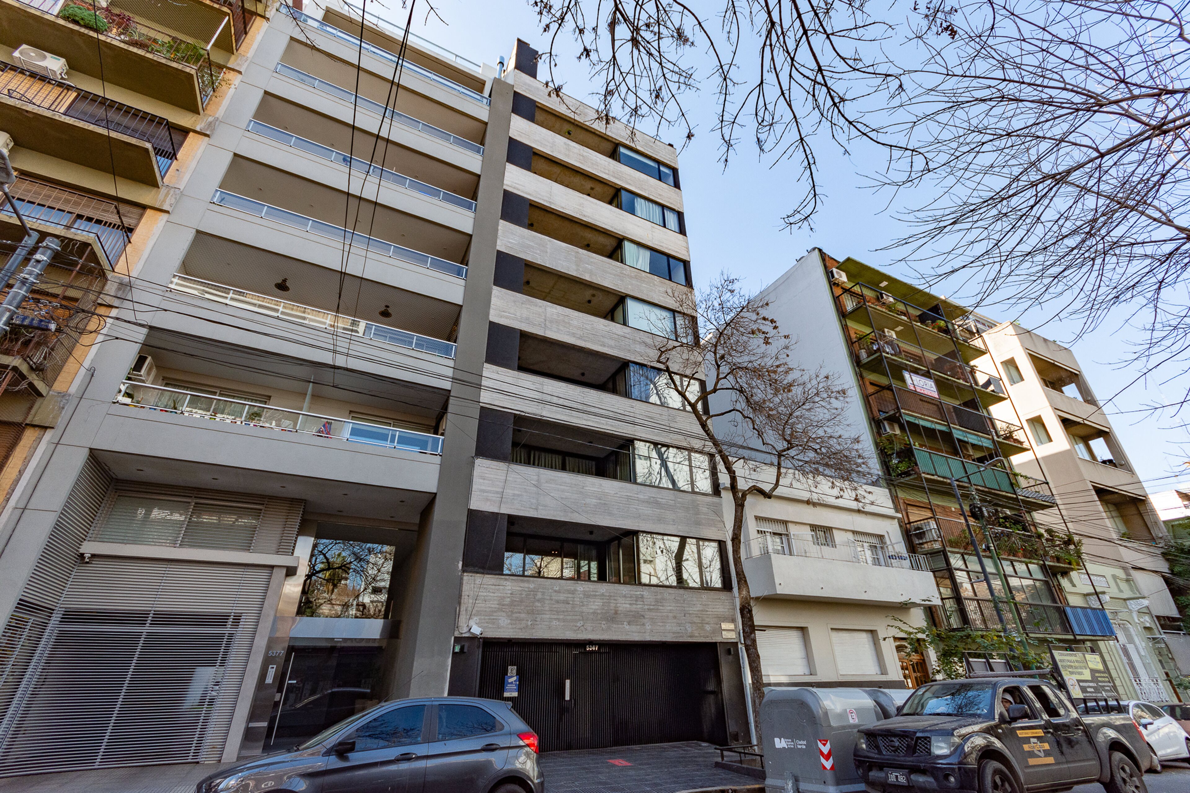 Un edificio de varios pisos con balcones, acceso a garaje y coches estacionados.