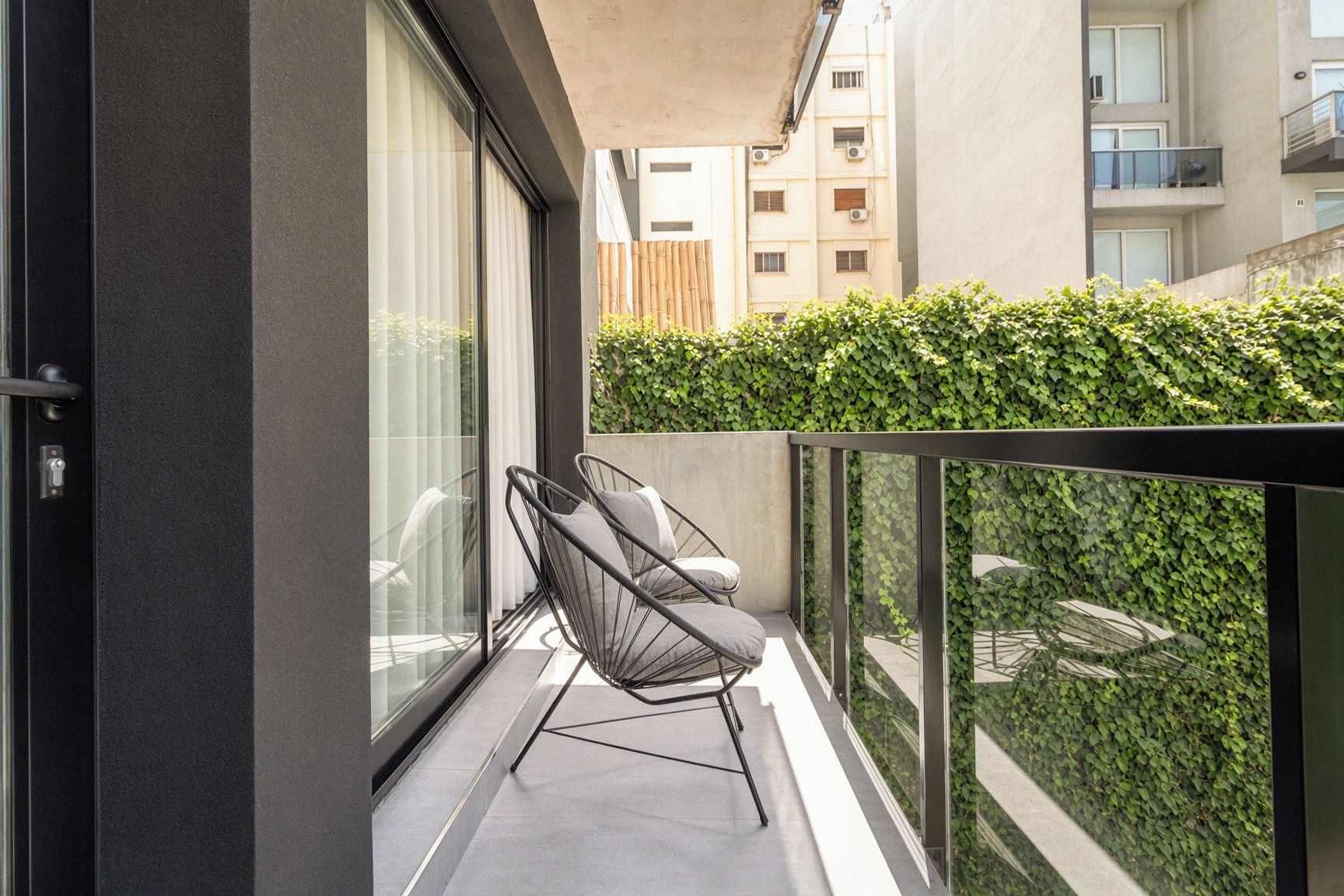 A balcony with a glass railing, two chairs, and a view of a building with a hedge.