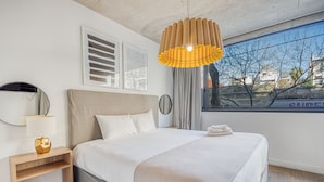 Una habitación de hotel moderna con una cama grande, una mesita de noche con lámpara, una ventana con vistas a árboles y una lámpara colgante original.