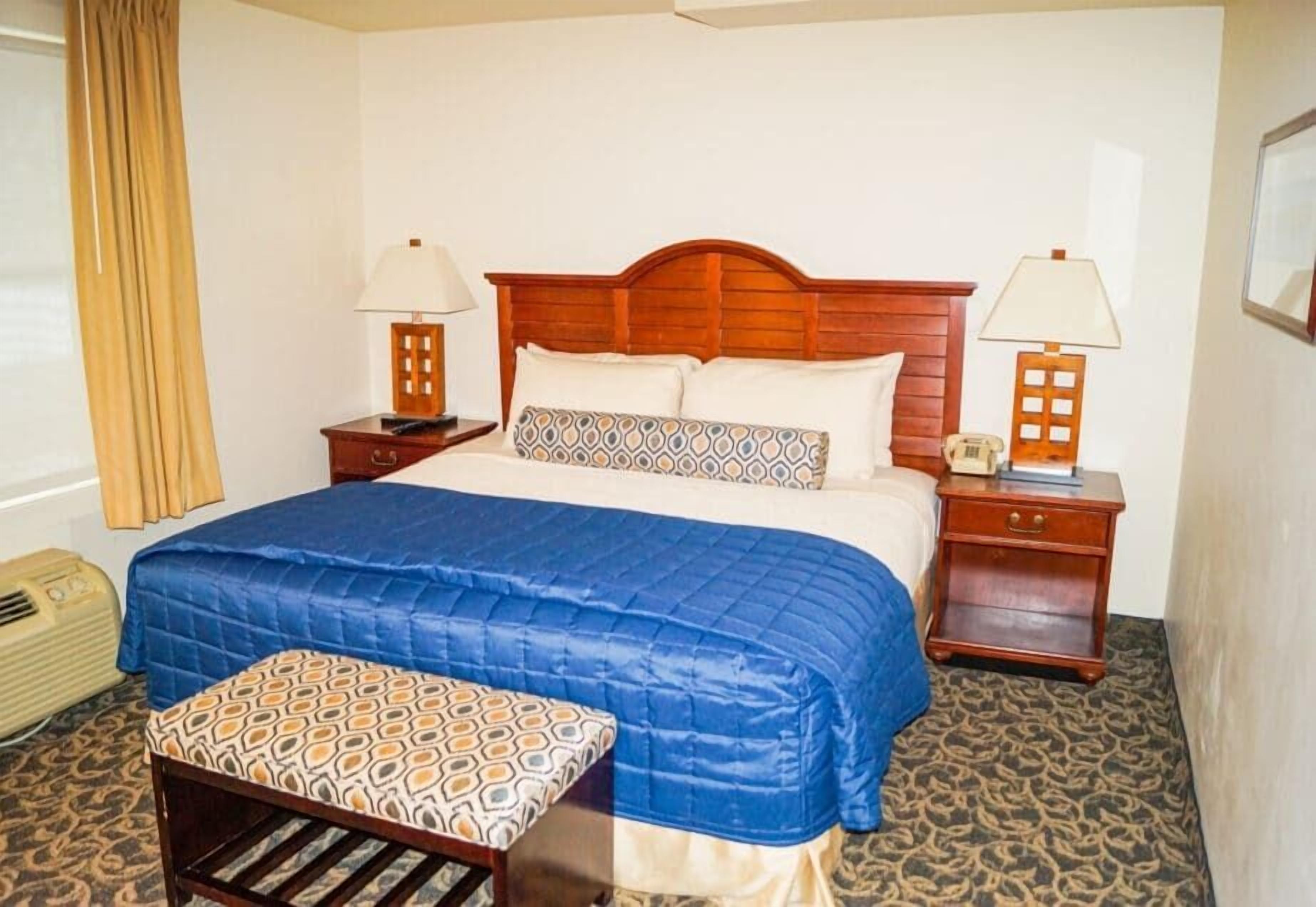 A hotel room with a wooden headboard bed, two bedside tables with lamps, a patterned bench, and a radiator.