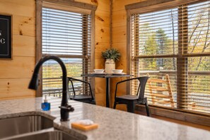 A kitchen with a sink, two chairs, a table with a potted plant, and large windows with blinds.