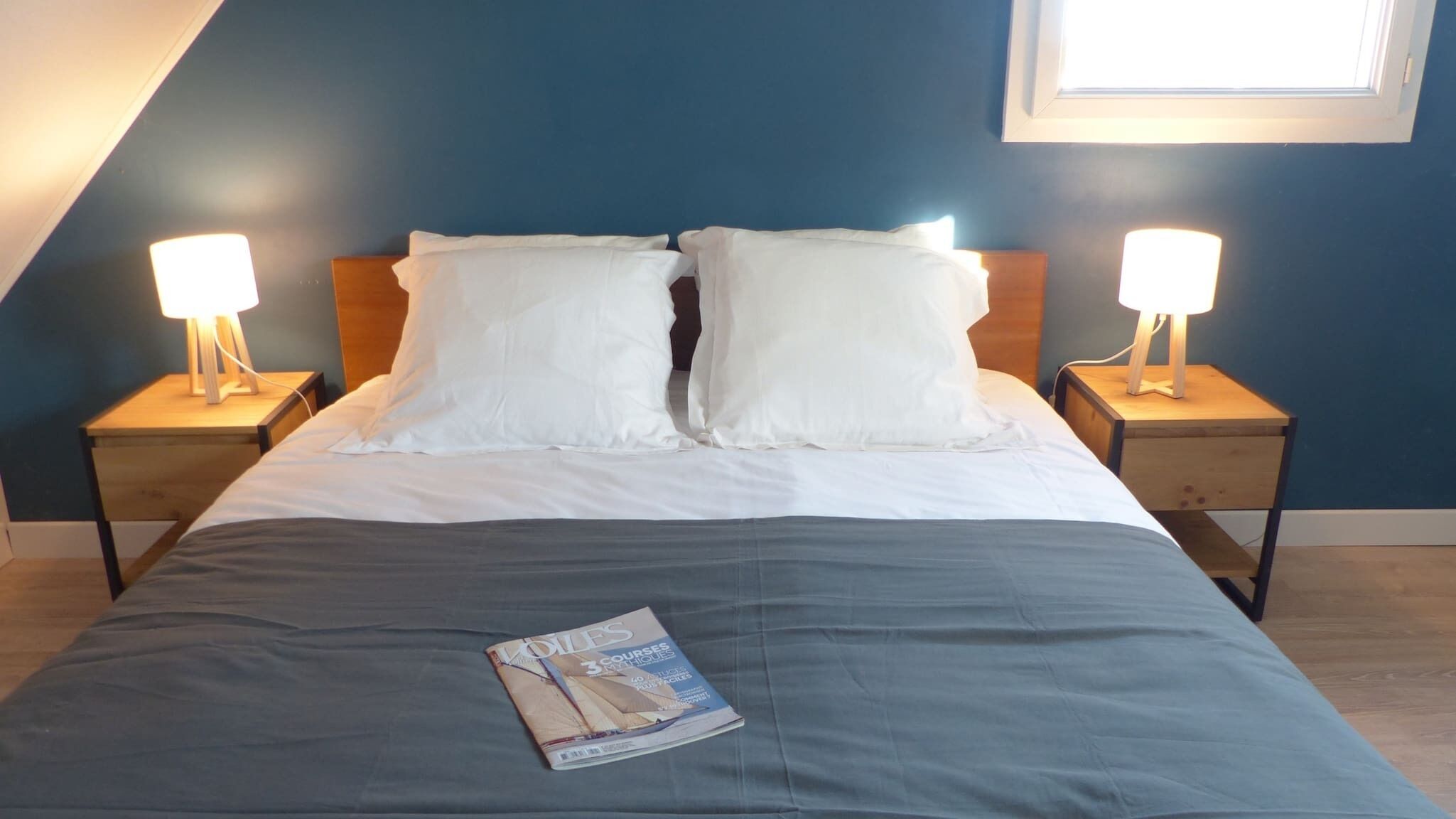 A neatly made bed with white linens, two bedside lamps, and a magazine on the bed.