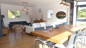 A modern living room with a wooden dining table, white chairs, a sofa, and a wicker chair. There are three pendant lights, a sailboat model, and a wall clock. - Comfortable 3* house with swimming pool (Larmor-Baden)