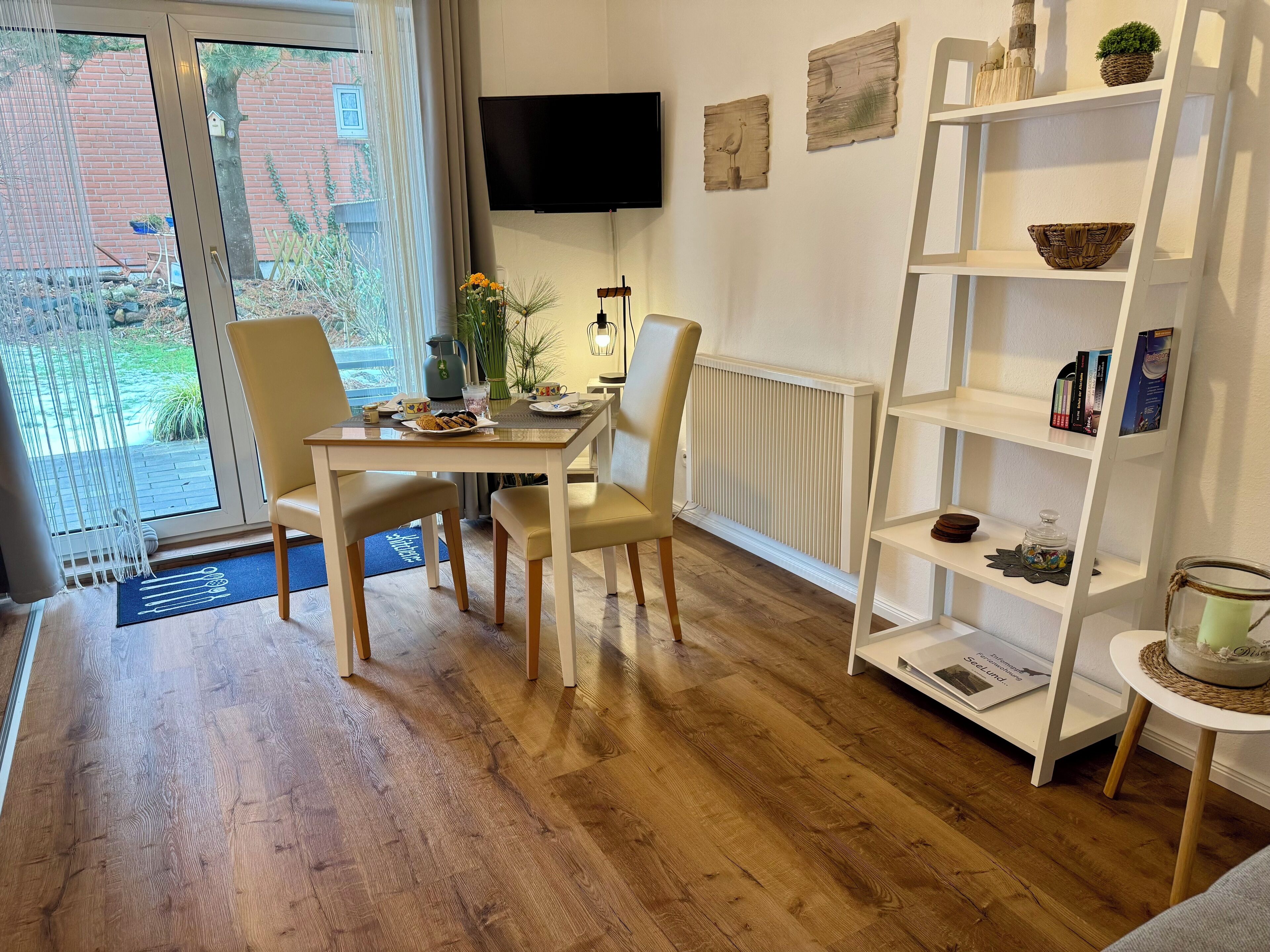 A modern dining area with a wooden table and chairs, a white bookshelf, and a television mounted on the wall.