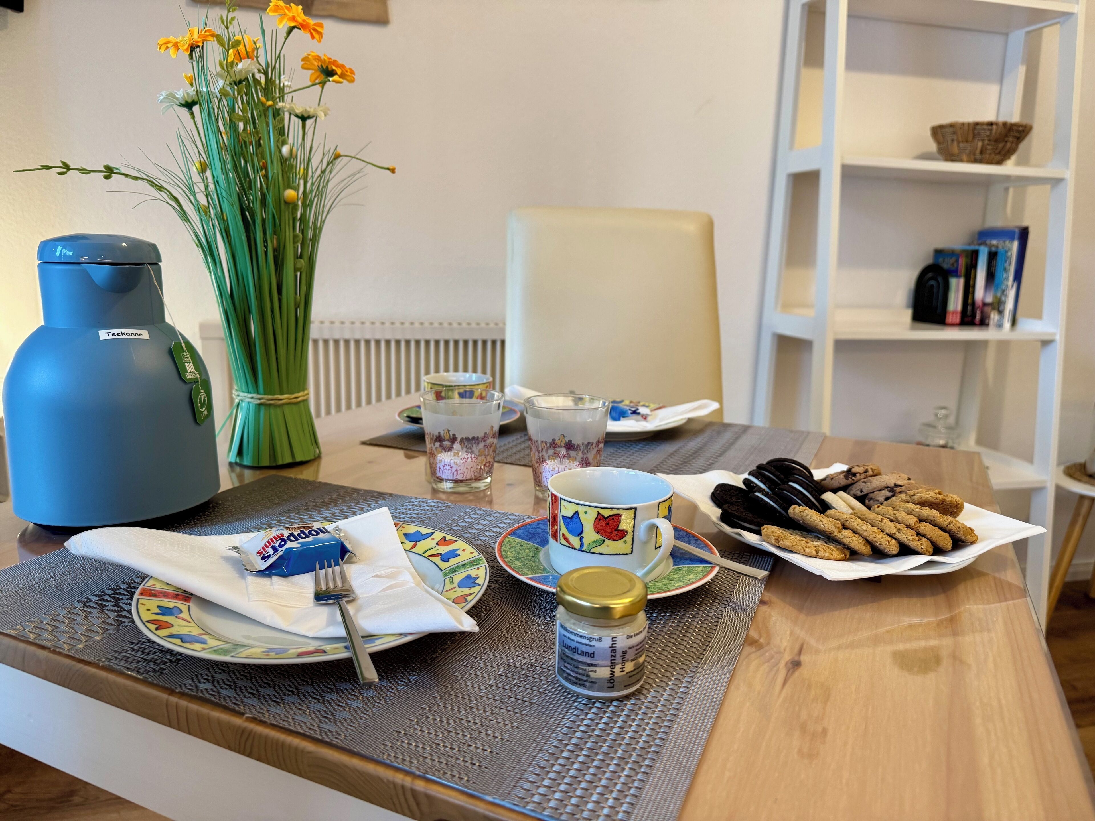 A dining table with a blue kettle, a vase with flowers, plates, glasses, and cookies.