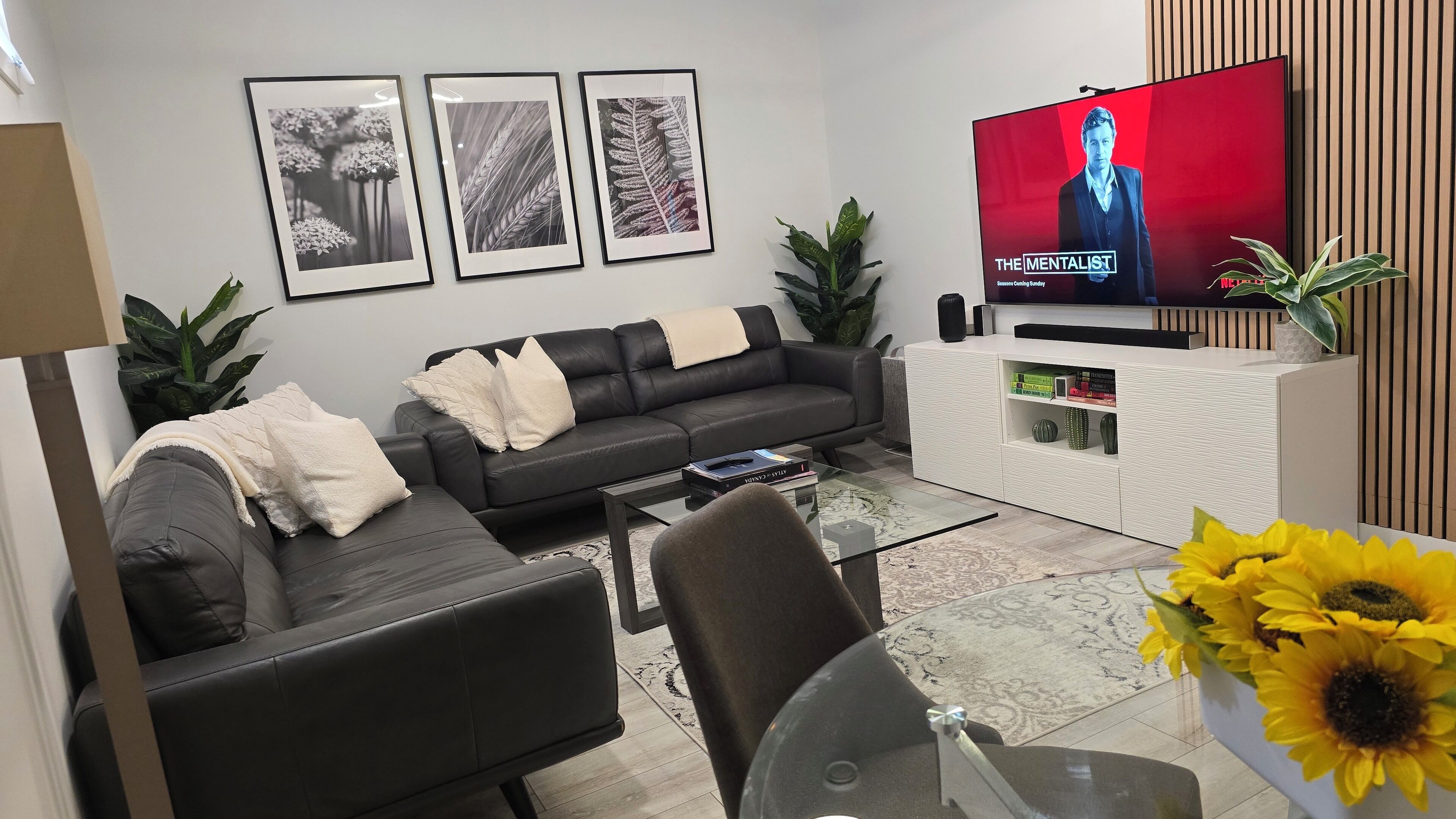 A modern living room with a black leather sofa, a glass coffee table, and a flat-screen TV displaying 'The Mentalist'. There are framed botanical prints on the wall, a white console table with decorative items, and a large potted plant.