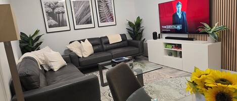 A modern living room with a black leather sofa, a glass coffee table, and a flat-screen TV displaying 'The Mentalist'. There are framed botanical prints on the wall, a white console table with decorative items, and a large potted plant.