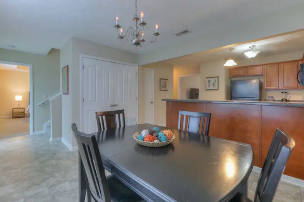 A dining area with a dark wooden table, chairs, and a chandelier. There is a kitchen area with wooden cabinets and a refrigerator.