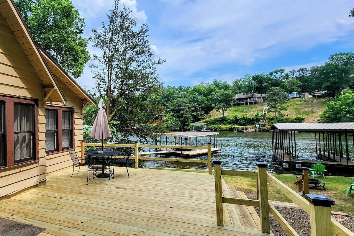 A wooden cabin with a deck overlooking a lake, featuring a table and chairs, a dock, and a covered area with chairs.