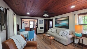 Ein Wohnzimmer mit braunem Ledersofa, einem beigen Sofa, einem Esstisch mit blauen Stühlen, einem Holztisch und einem Deckenventilator.