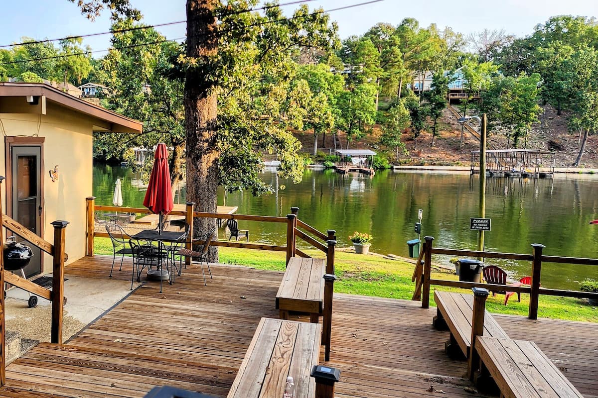 A wooden deck with outdoor seating, a small building with a door, and a view of a lake.