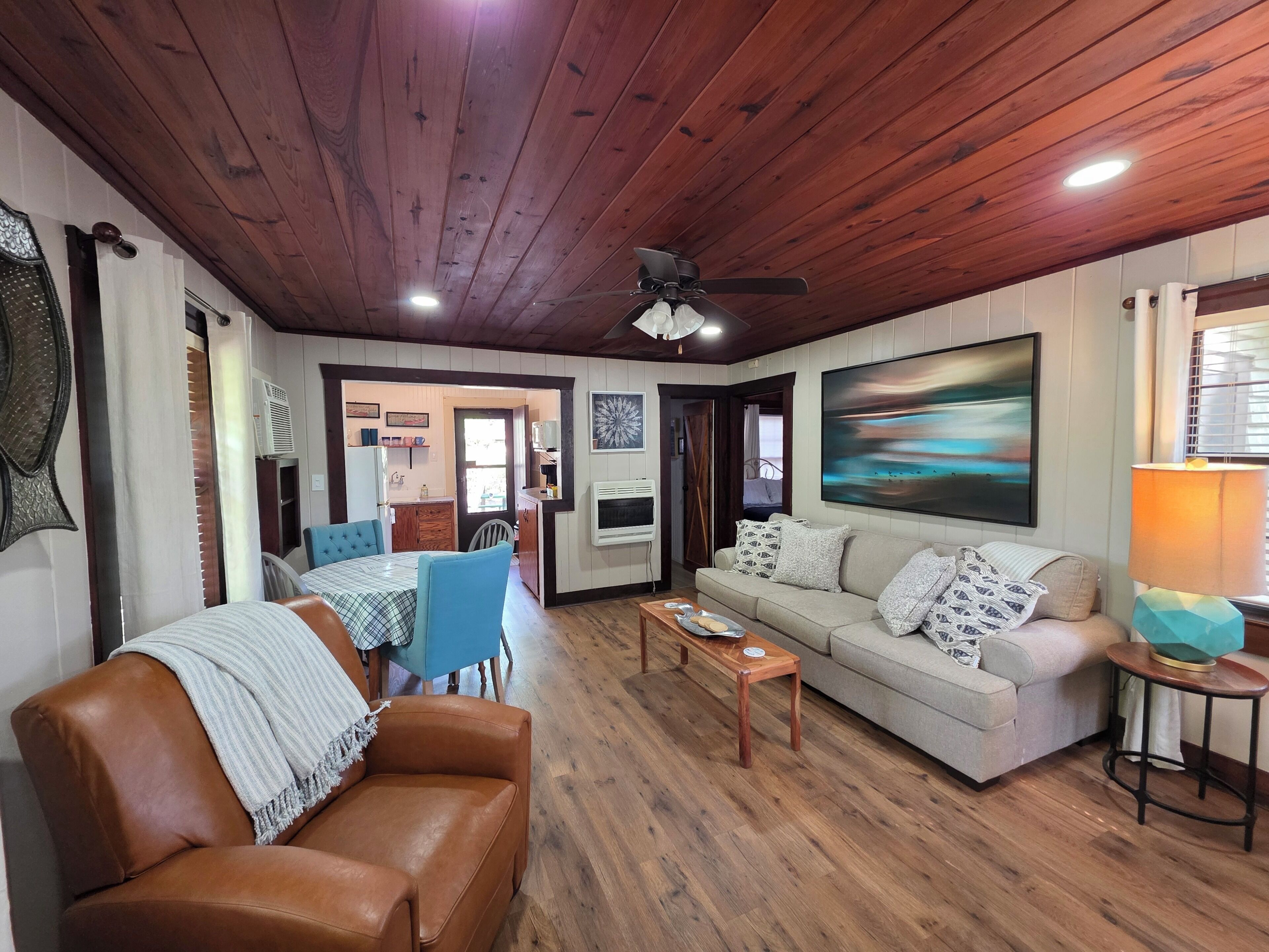 A living room with a brown leather sofa, a wooden coffee table, a beige sofa with patterned cushions, a dining area with a blue table and chairs, and a ceiling fan.