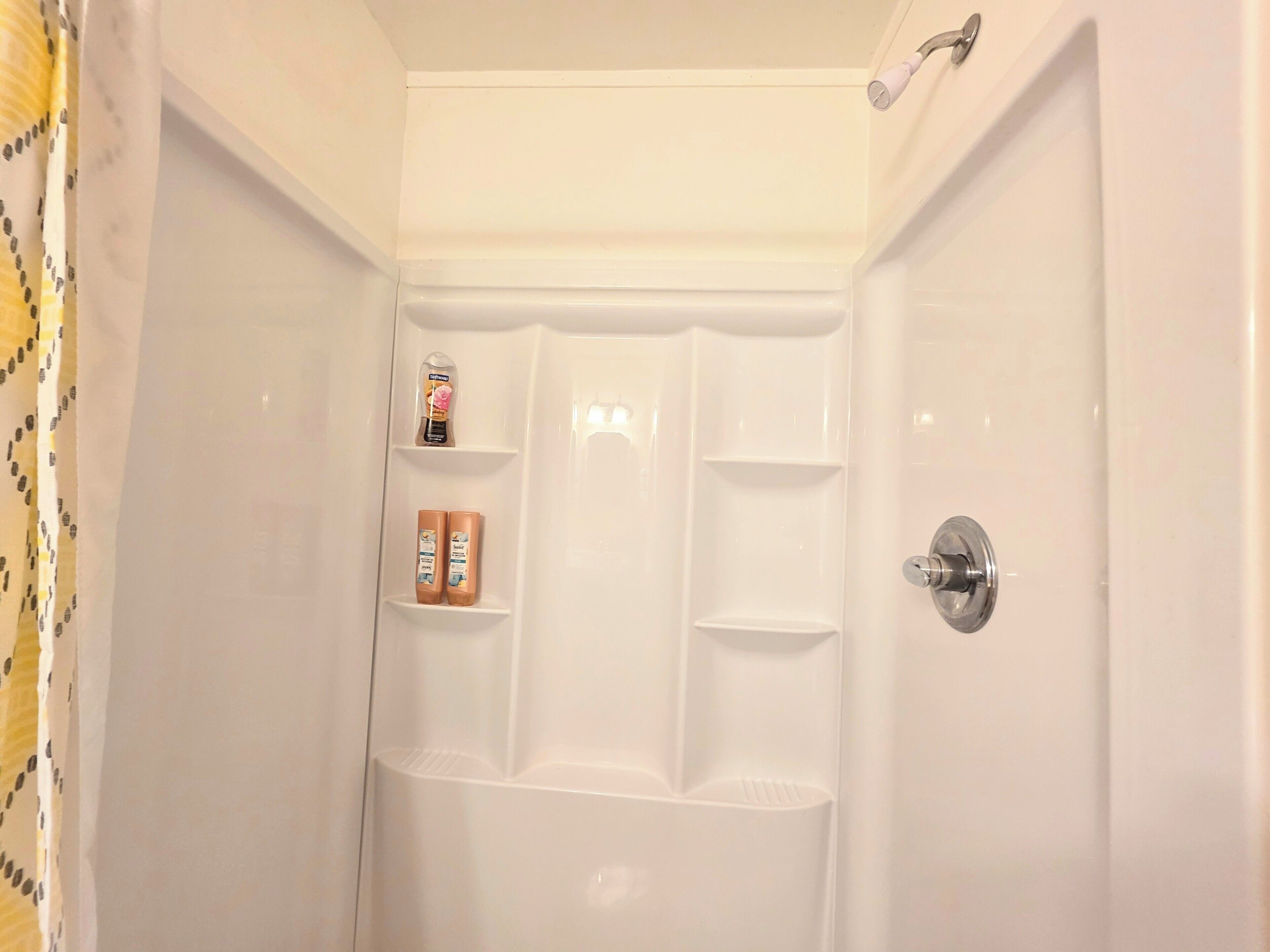 Cabina de ducha blanca con cabezal, dispensador de jabón y botella de champú en la pared.