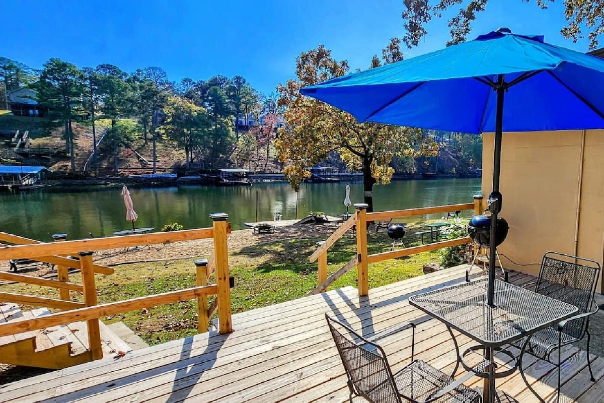 A wooden deck with a table and chairs, a blue umbrella, and a view of a lake with trees and a building in the background.
