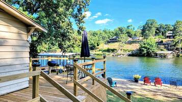 A wooden deck with outdoor furniture, a grill, and a view of a lake with houses in the background.