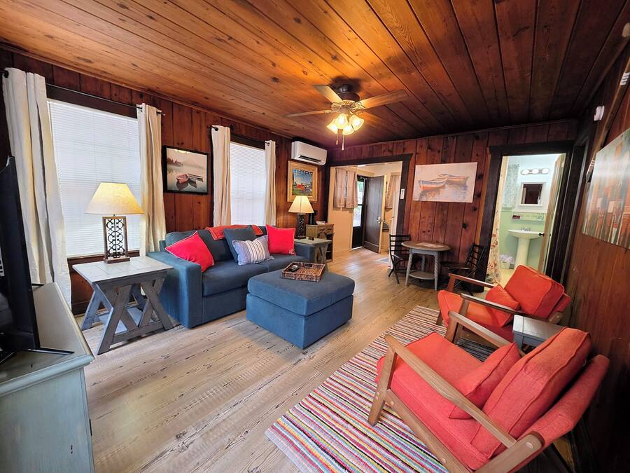 A cozy room with a wooden ceiling, a sofa with red cushions, a blue ottoman, a small table, and a colorful striped rug.