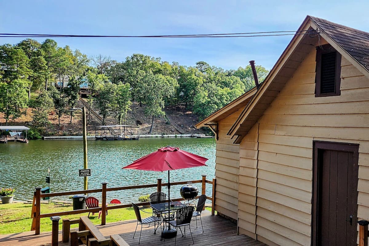 A cabin with a deck overlooking a lake, a red umbrella, and a grill.