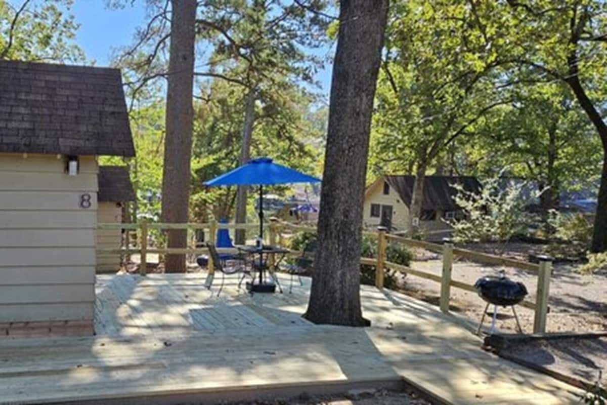 A wooden cabin with a deck, a blue umbrella, and a barbecue grill.