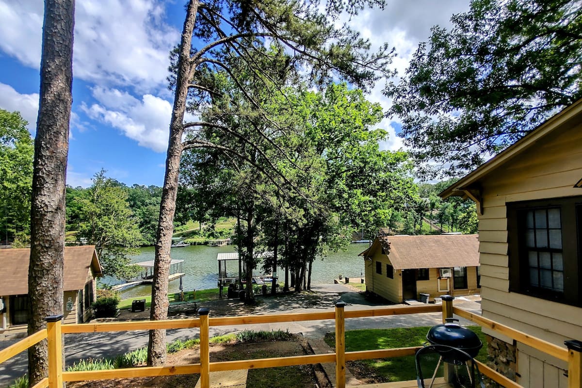 A wooden deck with a grill, overlooking a lake with docked boats and small cabins.