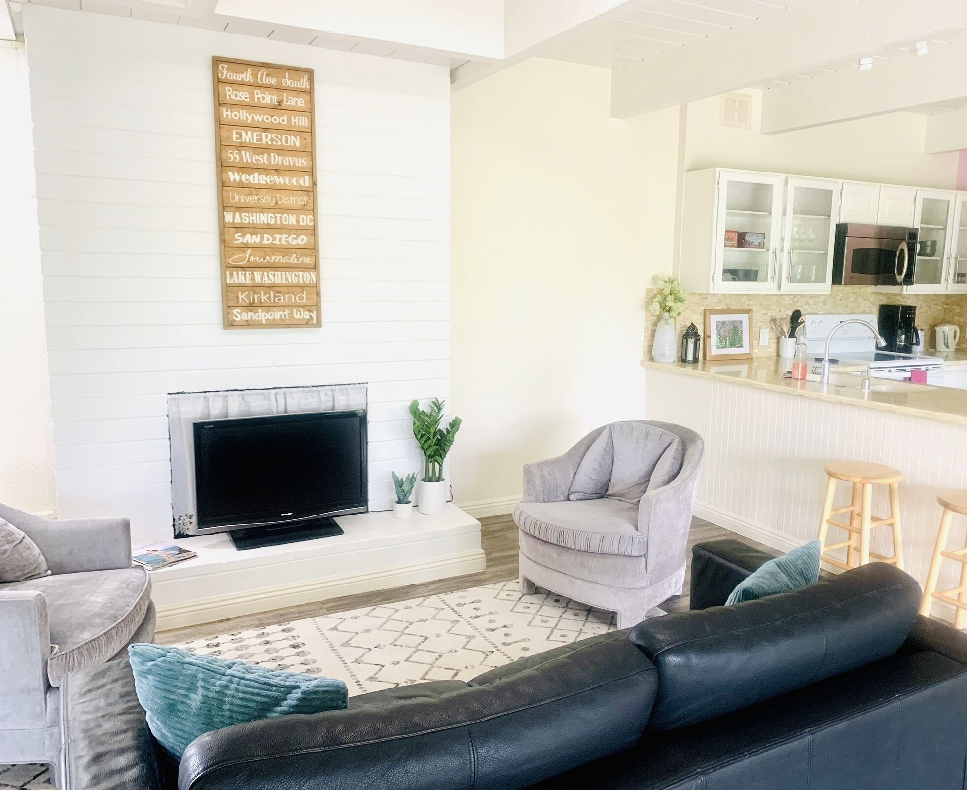 Una sala de estar con televisor de pantalla plana, chimenea, un sillón gris, un sofá de cuero negro y una zona de cocina con armarios y microondas.