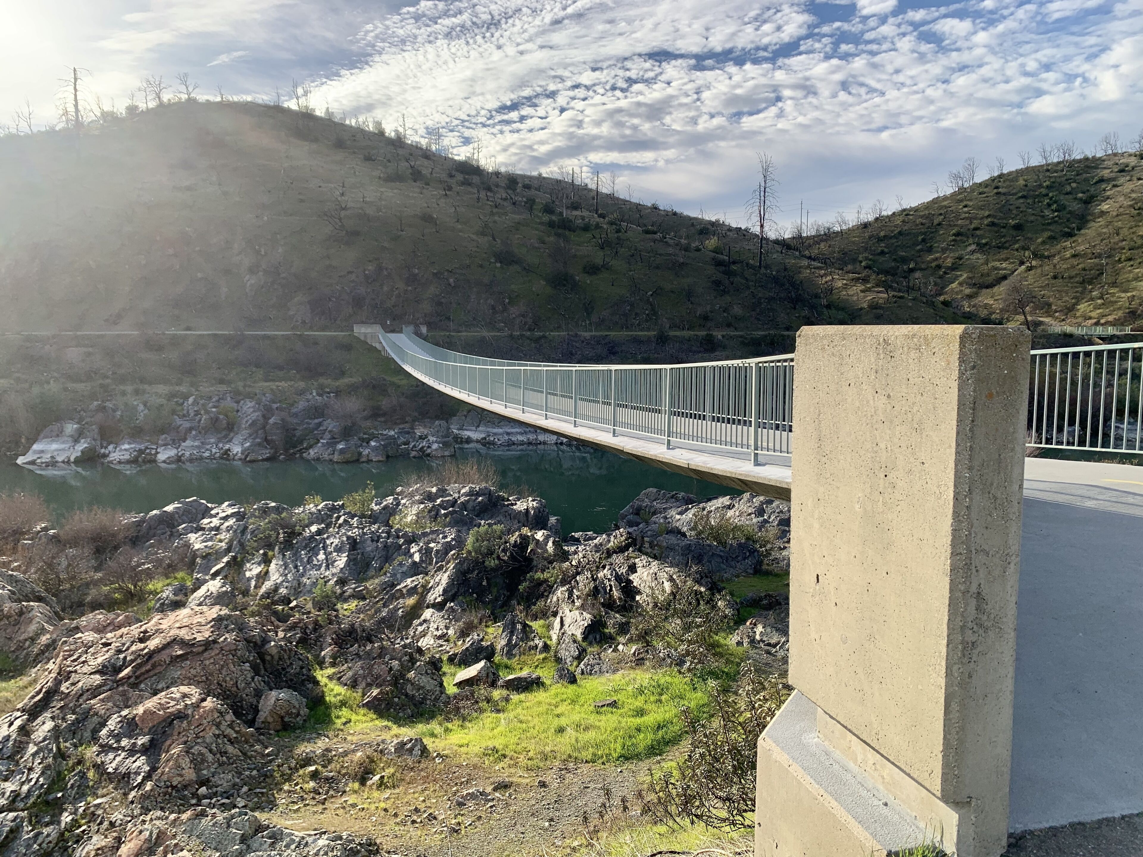 Een betonnen brug over een rivier, met een rotsachtige voorgrond en een heuvellandschap op de achtergrond.