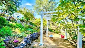 Um jardim com pérgola branca, muros de pedra e diversas plantas.