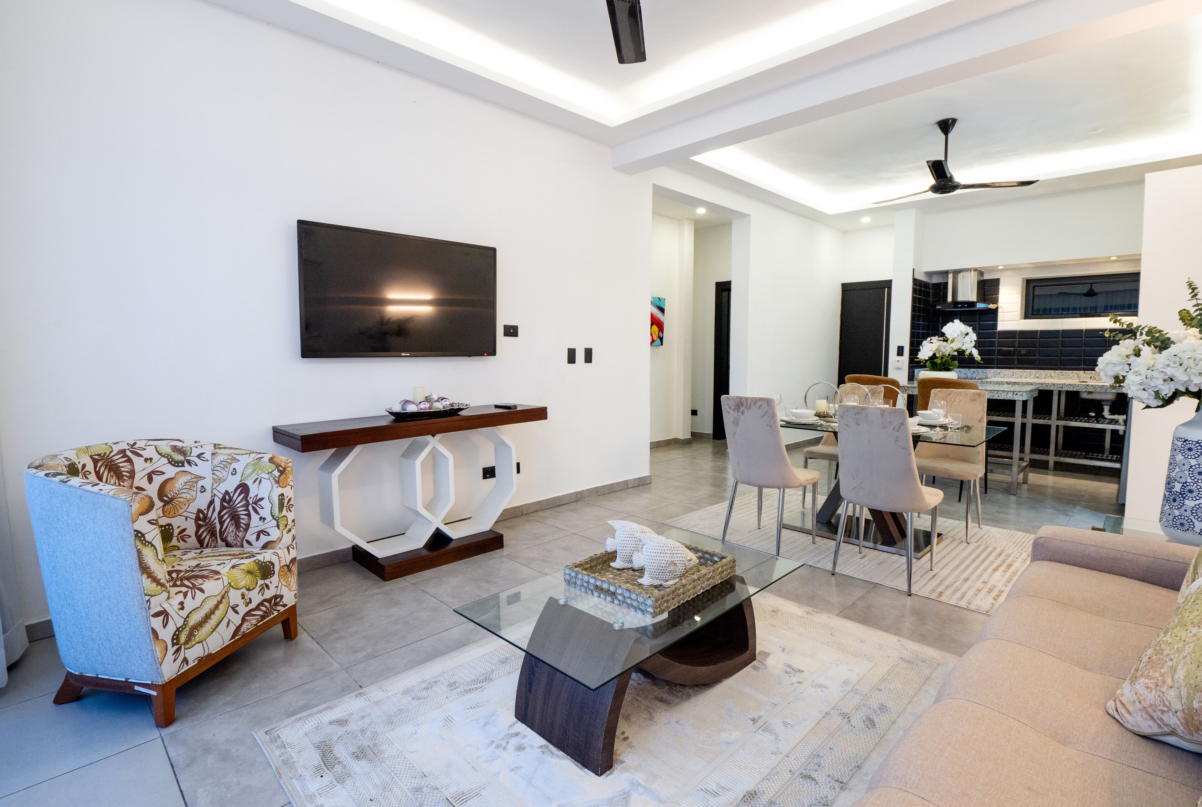 Een moderne woonkamer met een glazen salontafel, een bank, een aan de muur bevestigde televisie en een eethoek met stoelen en een tafel.