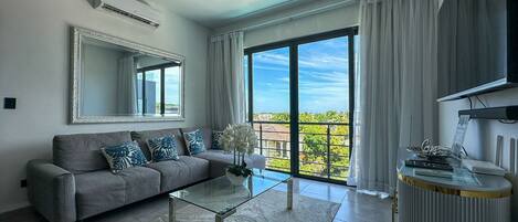 Un salón moderno con un sofá gris, una mesa de centro de cristal, un espejo grande y vistas a un balcón con un paisaje urbano.