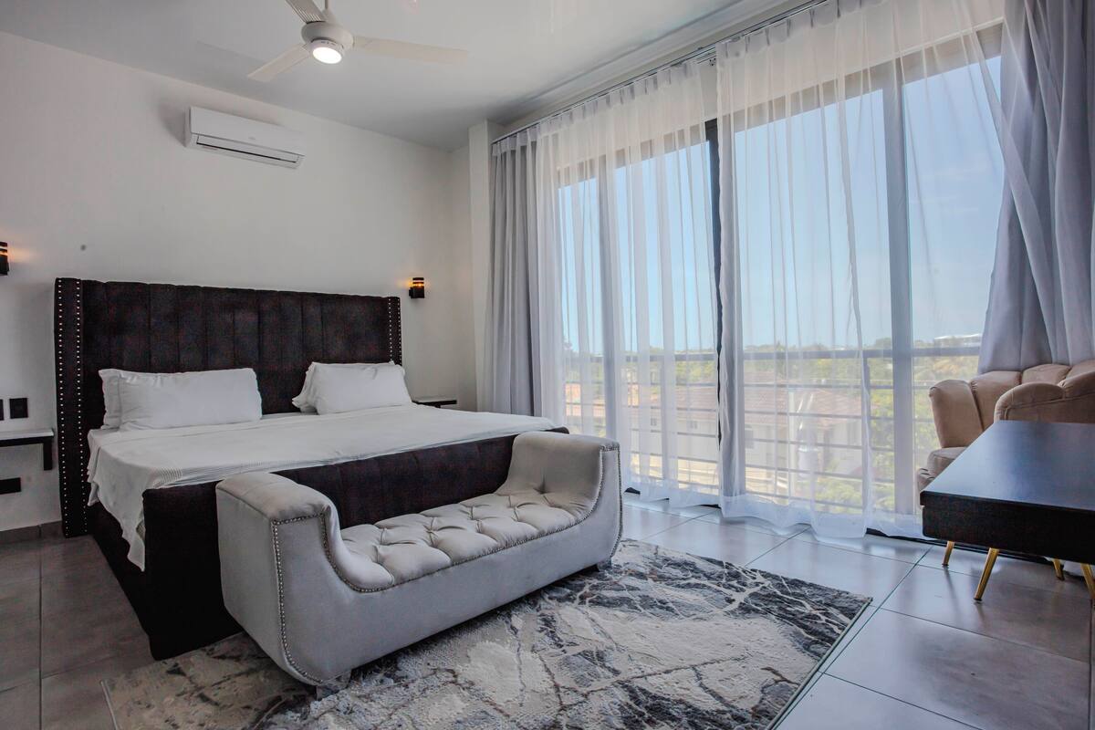 A modern bedroom with a large bed, a sofa, a balcony view, and a ceiling fan.