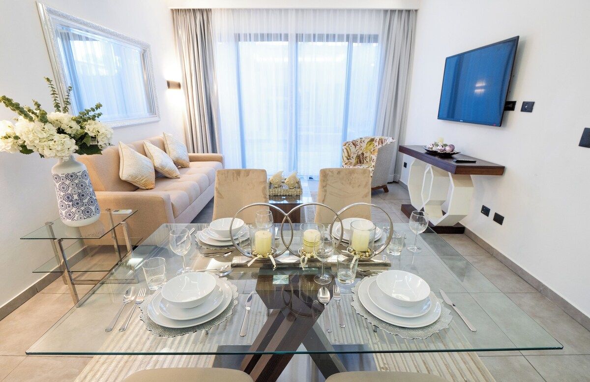 A dining area with a glass table set for four, surrounded by sofas and a TV mounted on the wall.