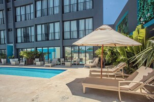 Zona de piscina con sillones de descanso y sombrillas, rodeada por un edificio con ventanas amplias y una pared azul.
