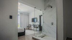 A modern bathroom with a glass-enclosed shower, a large mirror, and a visible bedroom with a bed and chairs.