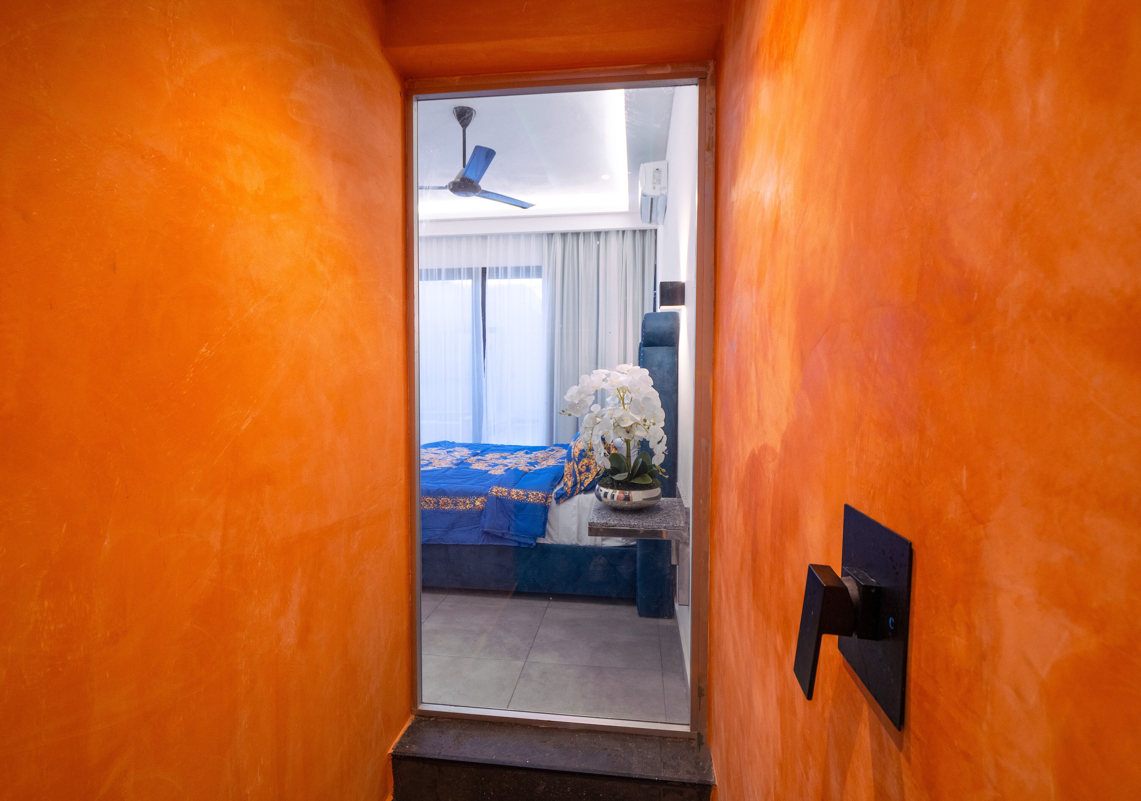 A narrow hallway leading to a bedroom with a bed, a nightstand, and a potted orchid.