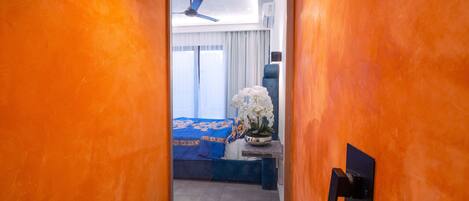A narrow hallway leading to a bedroom with a bed, a nightstand, and a potted orchid.