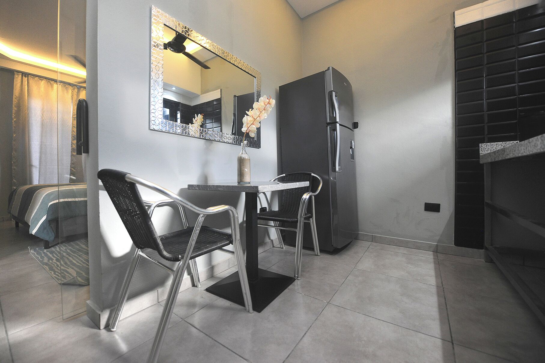 A compact room with a bed, a dining table with chairs, a refrigerator, and a mirror with a decorative light fixture.