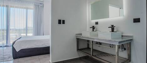 A modern hotel room with a large bed, a bathroom with a white sink and mirror, and a view through a large window.
