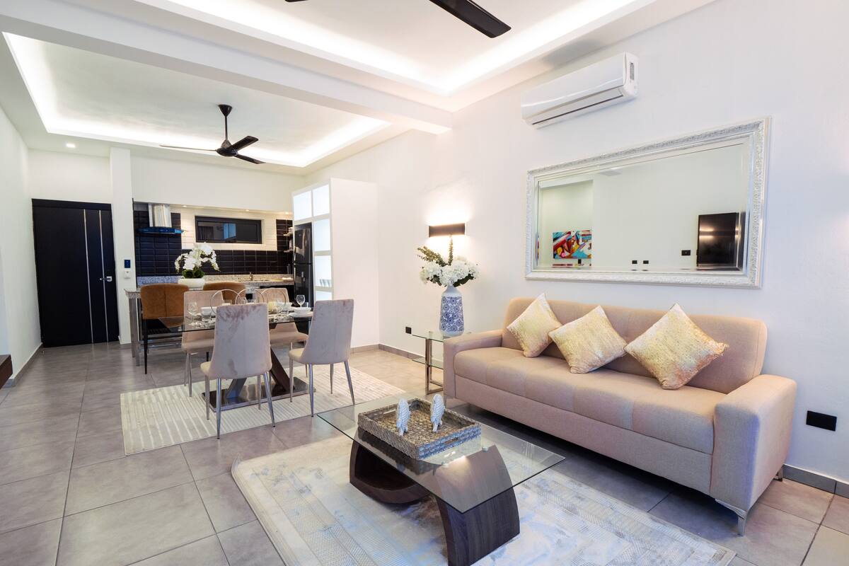 A modern living room with a glass coffee table, a beige sofa, and a dining area with chairs and a table.